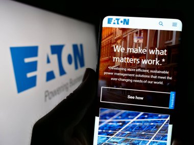 Stuttgart, Germany - 07-01-2023: Person holding cellphone with webpage of power management company Eaton Corporation plc on screen with logo. Focus on center of phone display. clipart