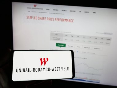 Stuttgart, Germany - 07-01-2023: Person holding mobile phone with logo of real estate company Unibail-Rodamco-Westfield SE on screen in front of web page. Focus on phone display. clipart