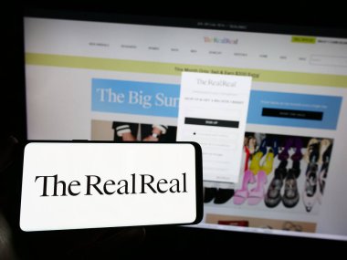 Stuttgart, Germany - 06-28-2023: Person holding smartphone with logo of US e-commerce company The RealReal Inc. on screen in front of website. Focus on phone display. clipart