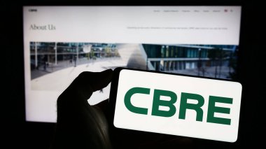 Stuttgart, Almanya - 06-25-2023: ABD 'li emlak şirketi CBRE Group Inc.' nin logosuyla cep telefonu sahibi kişi iş sayfasının önünde ekranda. Telefon ekranına odaklan.