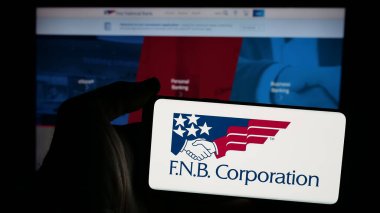 Stuttgart, Germany - 11-13-2024: Person holding mobile phone with logo of US company FNB Corporation (First National Bank) on screen in front of web page. Focus on phone display. clipart