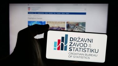 Stuttgart, Germany - 11-22-2024: Person holding mobile phone with logo of Croatian Bureau of Statistics (DZS) on screen in front of web page. Focus on phone display. clipart