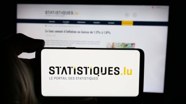 Stuttgart, Germany - 11-24-2024: Person holding cellphone with logo of Luxembourgish statistics agency STATEC on screen in front of webpage. Focus on phone display. clipart