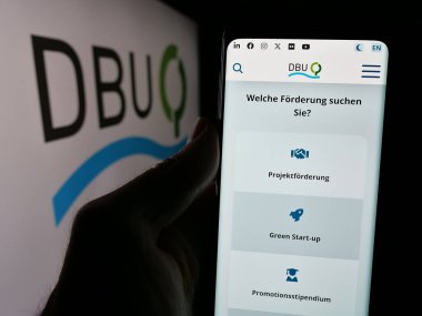 Stuttgart, Almanya - 11-20-2024: Broşürde Deutsche Bundesstiftung Umwelt (DBU) vakfının web sayfası bulunan kişi var. Telefon ekranının merkezine odaklan.