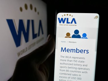 Stuttgart, Germany - 11-20-2024: Person holding cellphone with webpage of organisation World Lottery Association (WLA) on screen in front of logo. Focus on center of phone display. clipart