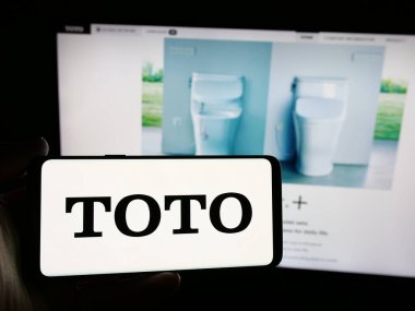 Stuttgart, Germany - 11-28-2024: Person holding smartphone with logo of Japanese toilet manufacturing company Toto Ltd. on screen in front of website. Focus on phone display. clipart