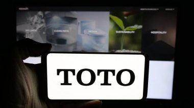 Stuttgart, Germany - 11-28-2024: Person holding cellphone with logo of Japanese toilet manufacturing company Toto Ltd. on screen in front of webpage. Focus on phone display. clipart