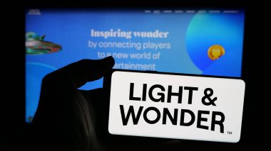 Stuttgart, Germany - 11-28-2024: Person holding mobile phone with logo of American gambling company Light and Wonder Inc. on screen in front of web page. Focus on phone display. clipart