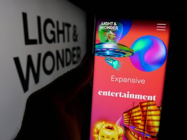 Stuttgart, Germany - 11-28-2024: Person holding smartphone with webpage of US gambling company Light and Wonder Inc. on screen in front of logo. Focus on center of phone display. clipart