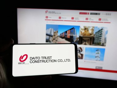 Stuttgart, Germany - 12-04-2024: Person holding smartphone with logo of company Daito Trust Construction Company Ltd. on screen in front of website. Focus on phone display. clipart