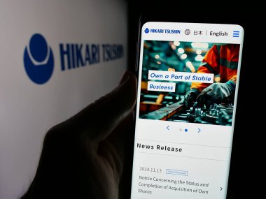 Stuttgart, Germany - 12-04-2024: Person holding cellphone with webpage of Japanese company Hikari Tsushin Inc. on screen in front of logo. Focus on center of phone display. clipart