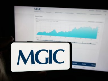 Stuttgart, Germany - 12-06-2024: Person holding smartphone with logo of US financial company MGIC Investment Corporation on screen in front of website. Focus on phone display. clipart