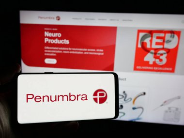 Stuttgart, Germany - 12-11-2024: Person holding smartphone with logo of US medical device company Penumbra Inc. on screen in front of website. Focus on phone display. clipart