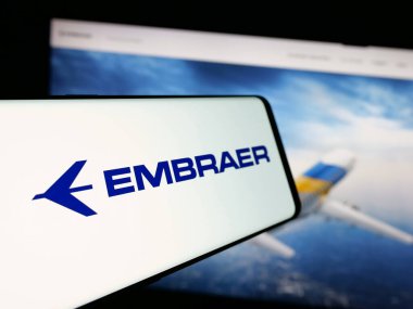 Stuttgart, Almanya - 12-16-2024: Brezilyalı havacılık şirketi Embraer SA 'nın logosunun yer aldığı cep telefonu iş sitesinin önünde ekranda. Telefon ekranının merkez soluna odaklan.