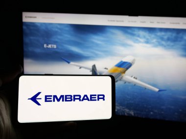 Stuttgart, Almanya - 12-16-2024: Brezilyalı havacılık şirketi Embraer SA 'nın logosunu elinde tutan kişi web sitesinin önünde ekranda. Telefon ekranına odaklan.