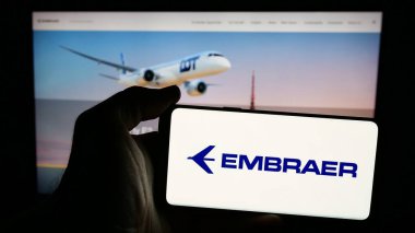 Stuttgart, Almanya - 12-16-2024: Brezilya havacılık şirketi Embraer SA 'nın logosunu taşıyan cep telefonu sahibi kişi iş sayfasının önünde ekranda. Telefon ekranına odaklan.