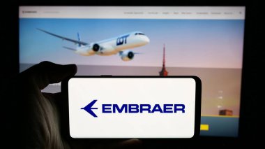 Stuttgart, Almanya - 12-16-2024: Brezilyalı havacılık şirketi Embraer SA 'nın logosuyla cep telefonu sahibi kişi iş sayfasının önünde ekranda. Telefon ekranına odaklan.