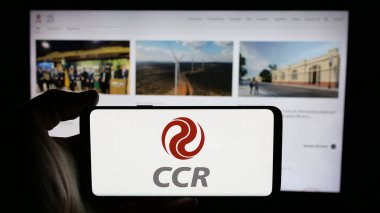 Stuttgart, Germany - 12-16-2024: Person holding cellphone with logo of company Companhia de Concessoes Rodoviarias SA (CCR) on screen in front of web page. Focus on phone display. clipart