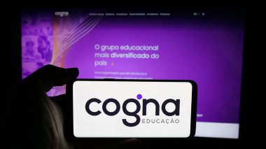 Stuttgart, Almanya - 12-18-2024: Brezilya eğitim şirketi Cogna Educacao SA 'nın logosuyla cep telefonu sahibi kişi iş sayfasının önünde ekranda görülüyor. Telefon ekranına odaklan.