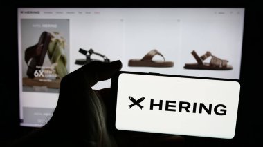 Stuttgart, Germany - 12-18-2024: Person holding mobile phone with logo of Brazilian fashion company Hering on screen in front of business web page. Focus on phone display. clipart