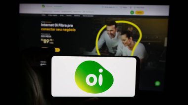 Stuttgart, Germany - 12-19-2024: Person holding cellphone with logo of Brazilian telecommunications company Oi SA on screen in front of business webpage. Focus on phone display. clipart