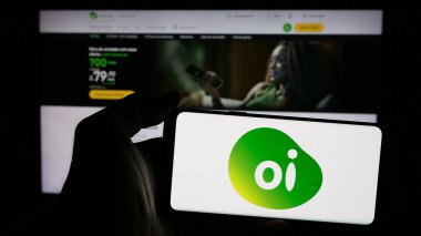 Stuttgart, Germany - 12-19-2024: Person holding mobile phone with logo of Brazilian telecommunications company Oi SA on screen in front of web page. Focus on phone display. clipart