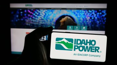 Stuttgart, Germany - 12-20-2024: Person holding mobile phone with logo of American utility company Idaho Power Company (IPC) on screen in front of web page. Focus on phone display. clipart