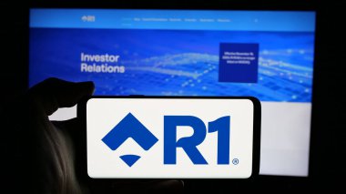 Stuttgart, Almanya - 12-20-2024: ABD 'li hasta deneyimi şirketi R1 RCM Inc.' nin logosuyla cep telefonu sahibi kişi iş sayfasının önünde ekranda. Telefon ekranına odaklan.