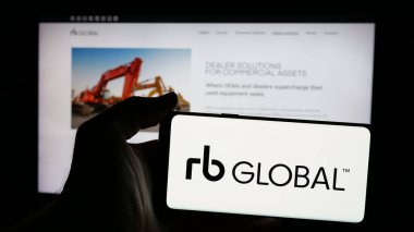 Stuttgart, Germany - 12-27-2024: Person holding mobile phone with logo of company RB Global Inc. (Ritchie Bros. Auctioneers) on screen in front of web page. Focus on phone display. clipart