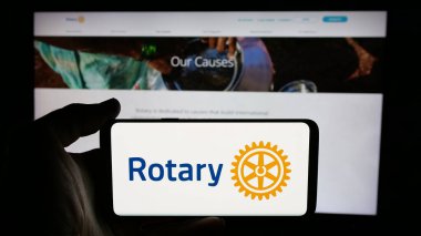 Stuttgart, Germany - 12-30-2024: Person holding cellphone with logo of service club Rotary International on screen in front of webpage. Focus on phone display. clipart