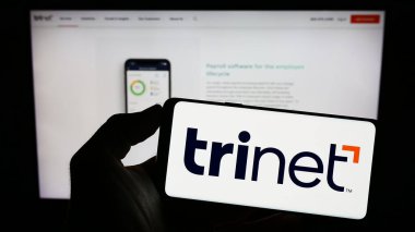 Stuttgart, Almanya - 06-18-2023: ABD 'li taşeronluk şirketi TriNet Group Inc.' in logosuyla cep telefonu sahibi kişi iş sayfasının önünde ekranda. Telefon ekranına odaklan.
