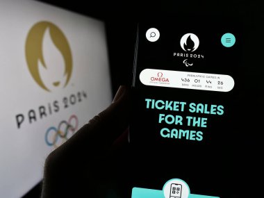 Stuttgart, Germany - 06-19-2023: Person holding cellphone with webpage of the 2024 Summer Olympics in Paris on screen in front of logo. Focus on center of phone display. clipart