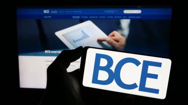 Stuttgart, Germany - 06-23-2023: Person holding cellphone with logo of Canadian telecommunications company BCE Inc. on screen in front of webpage. Focus on phone display. clipart