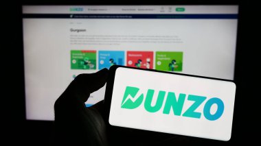 Stuttgart, Almanya - 04-23-2023: Hintli dağıtım uygulaması şirketi Dunzo Digital 'in logosuyla cep telefonu sahibi kişi iş sayfasının önünde ekranda. Telefon ekranına odaklan.