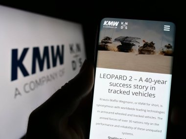 Stuttgart, Almanya - 04-25-2023: Cep telefonunda Krauss-Maffei Wegmann GmbH Co. KG (KMW) sitesi bulunan kişi logonun önünde ekranda. Telefon ekranının merkezine odaklan.