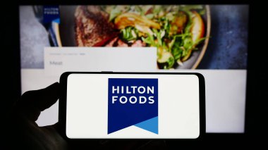 Stuttgart, Almanya - 04-25-2023: İngiliz Hilton Food Group plc şirketinin logosuna sahip akıllı telefon sahibi kişi web sitesinin önünde ekranda. Telefon ekranına odaklan.