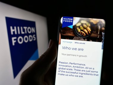 Stuttgart, Germany - 04-25-2023: Person holding cellphone with webpage of British company Hilton Food Group plc on screen in front of logo. Focus on center of phone display. clipart