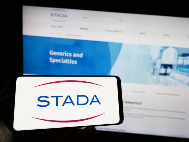 Stuttgart, Germany - 04-25-2023: Person holding mobile phone with logo of German company Stada Arzneimittel AG on screen in front of business web page. Focus on phone display. clipart