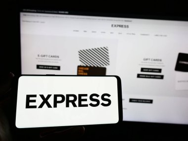 Stuttgart, Germany - 04-25-2023: Person holding smartphone with logo of US fashion retail company Express Inc. on screen in front of website. Focus on phone display. clipart