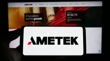 Stuttgart, Germany - 04-27-2023: Person holding cellphone with logo of US electronics company AMETEK Inc. on screen in front of business webpage. Focus on phone display. clipart