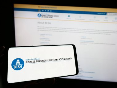 Stuttgart, Almanya - 04-28-2023: İnternet sitesi önünde üzerinde İş logosu, Tüketici Hizmetleri ve Konut Hizmetleri olan akıllı telefon tutan kişi. Telefon ekranına odaklan.