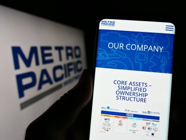 Stuttgart, Germany - 04-28-2023: Person holding cellphone with website of Metro Pacific Investments Corporation (MPIC) on screen with logo. Focus on center of phone display. clipart