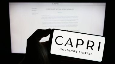 Stuttgart, Almanya - 06-17-2023: Moda şirketi Capri Holding Limited 'in logosuna sahip akıllı telefon sahibi kişi web sitesinin önünde ekranda. Telefon ekranına odaklan.