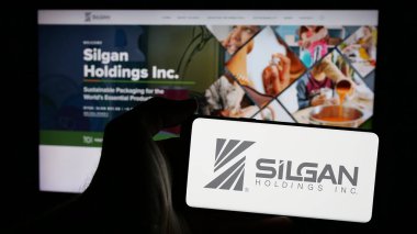 Stuttgart, Germany - 01-08-2024: Person holding mobile phone with logo of American packaging company Silgan Holdings Inc. on screen in front of web page. Focus on phone display. clipart