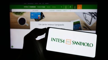 Stuttgart, Almanya - 04-16-2023: İtalyan bankacılık şirketi Intesa Sanpaolo SpA 'nın logosuyla cep telefonu tutan kişi iş sayfasının önünde ekranda. Telefon ekranına odaklan.