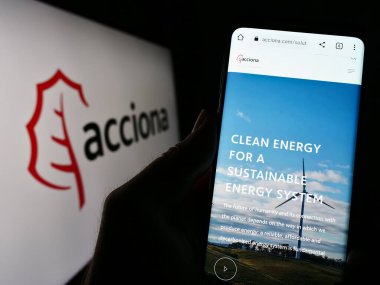 Stuttgart, Germany - 04-16-2023: Person holding cellphone with webpage of Spanish infrastructure company Acciona SA on screen in front of logo. Focus on center of phone display. clipart