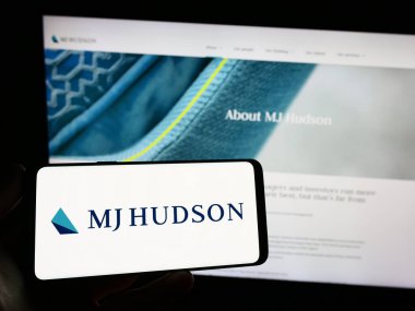 Stuttgart, Almanya - 04-19-2023: İngiliz finans şirketi MJ Hudson Group plc 'in logosuna sahip akıllı telefon sahibi kişi web sitesinin önünde ekranda. Telefon ekranına odaklan.