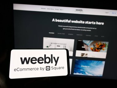 Stuttgart, Almanya - 04-19-2023: Web sunuculuğu şirketi Weebly 'nin logosuna sahip akıllı telefon sahibi kişi web sitesinin önünde ekranda. Telefon ekranına odaklan.