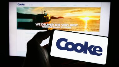 Stuttgart, Germany - 04-19-2023: Person holding mobile phone with logo of Canadian seafood company Cooke Aquaculture Inc. on screen in front of web page. Focus on phone display. clipart