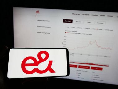 Stuttgart, Germany - 04-23-2023: Person holding mobile phone with logo of Emirates Telecommunications Group (etisalat) on screen in front of web page. Focus on phone display. clipart